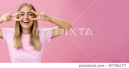 Joyful friendly and peaceful common european woman with blond hair in casual t-shirt showing peace or disco signs over eyes smiling cheerfully at camera having fun against pink background 97796277
