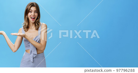 Woman presenting product over copy space. Good-looking charismatic adult tall woman with chestnut hair pointing at left side of wall and smiling broadly as if advertising against blue background 97796288