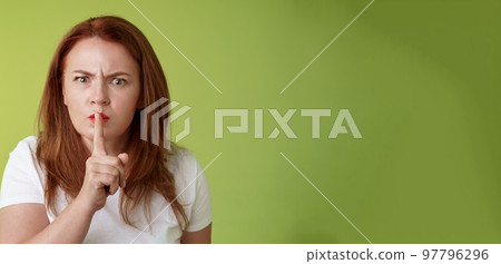 Not speak during exam. Strict serious-looking displeased middle-aged redhead woman frowning disappointed hushing say shush index finger pressed lips keep quiet gesture green background 97796296
