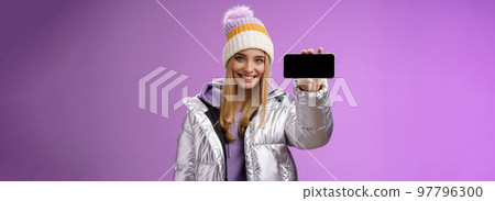 Confident friendly good-looking blond girl in outdoor silver glittering jacket hat hold smartphone horizontal showing mobile phone display assertive smile recommend use app, purple background Confident friendly good-looking blond girl in outdoor silver glittering jacket hat hold smartphone horizontal showing mobile phone display assertive smile recommend use app, purple background 97796300