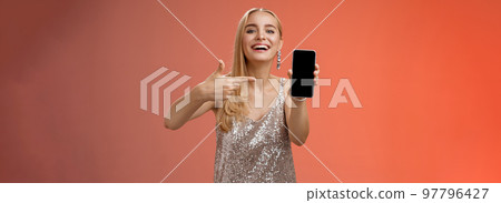 Boastful proud charming elegant blond woman in stylish evening dress show smartphone display proudly pointing mobile phone screen smiling showing photo boyfriend, standing red background 97796427