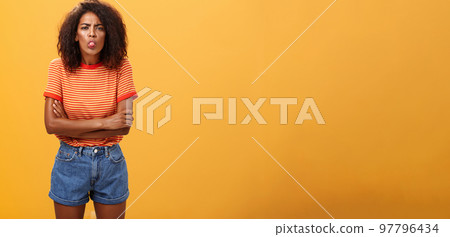 Immature girl showing bad side of character. Portrait of childish offended or displeased young African-American woman with curly hair showing tongue crossing arms on chest over orange background Immature girl showing bad side of character. Portrait of childish offended or displeased young African-American woman with curly hair showing tongue crossing arms on chest over orange background 97796434