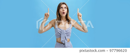 Waist-up shot of curious good-looking stylish modern woman dropping jaw saying wow while lifting head up looking and pointing upwards with amazed, surprised expression posing against blue background Waist-up shot of curious good-looking stylish modern woman dropping jaw saying wow while lifting head up looking and pointing upwards with amazed, surprised expression posing against blue background 97796620