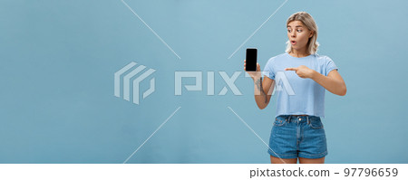 You should download app. Portrait of amazed shocked attractive blonde girl in trendy summer t-shirt holding smartphone gasping from surprise pointing at gadget screen posing over blue background 97796659
