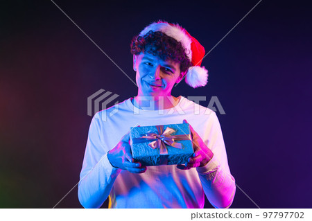Man with curly hair gives gift box by hands to camera on gradient neon background. Santa guy smiling 97797702