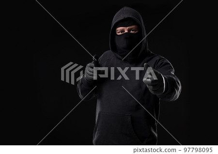 Masked robber with gun aiming into the camera against a black background Masked robber with gun aiming into the camera against a black background 97798095
