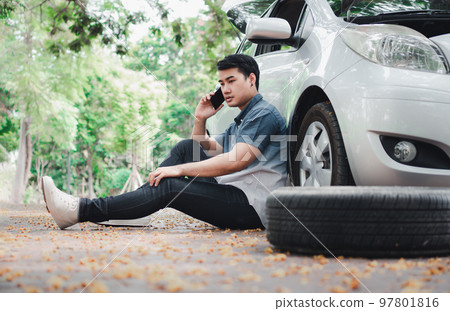Asian man sitting beside car and using mobile phone calling for assistance after a car breakdown on street. Concept of vehicle engine problem or accident and emergency help from Professional mechanic 97801816