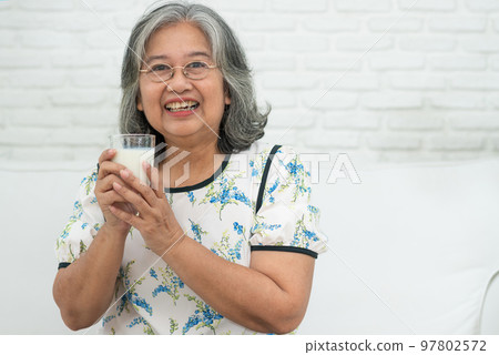 Asian Senior woman holding glass milk while relaxing on a sofa living room for retirement wellness. Elderly Woman drinking a glass of milk to maintain her wellbeing. Concept of wellness and healthy. Asian Senior woman holding glass milk while relaxing on a sofa living room for retirement wellness. Elderly Woman drinking a glass of milk to maintain her wellbeing. Concept of wellness and healthy. 97802572