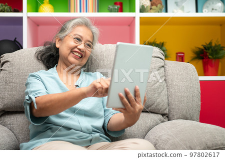Senior asian woman wearing glasses and relaxing at home on a sofa and using tablet for reading news and e-book. Concept of technology for elderly. 97802617