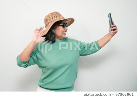 Portrait of happy Asian senior woman wearing sunglasses, a hat, and holding smartphone for a selfie, standing isolated on white background. Concept of tourist enjoy and fun valentine day after retire Portrait of happy Asian senior woman wearing sunglasses, a hat, and holding smartphone for a selfie, standing isolated on white background. Concept of tourist enjoy and fun valentine day after retire 97802961