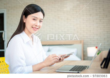 Happy beautiful asian woman working on a laptop and smartphone at the home office sitting at table. Happy female professional freelancer online using notebook pc and smartphone concept. Happy beautiful asian woman working on a laptop and smartphone at the home office sitting at table. Happy female professional freelancer online using notebook pc and smartphone concept. 97803063