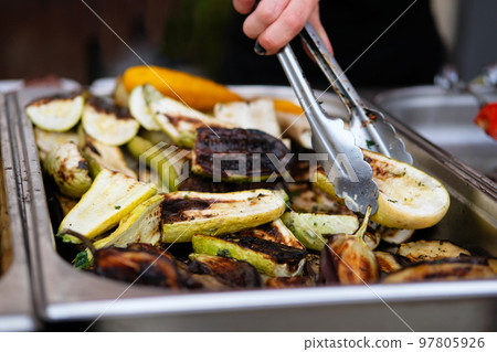 Grilled vegetables on open kitchen food festival event. Street food and outdoor cooking concept. Hand of woman with thongs. 97805926