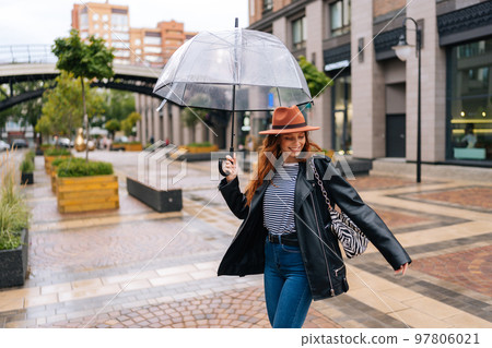 Happy excited young woman in elegant hat dancing and having fun with transparent umbrella on beautiful city street, enjoying rainy weather outdoors. Concept of female lifestyle at autumn season. Happy excited young woman in elegant hat dancing and having fun with transparent umbrella on beautiful city street, enjoying rainy weather outdoors. Concept of female lifestyle at autumn season. 97806021