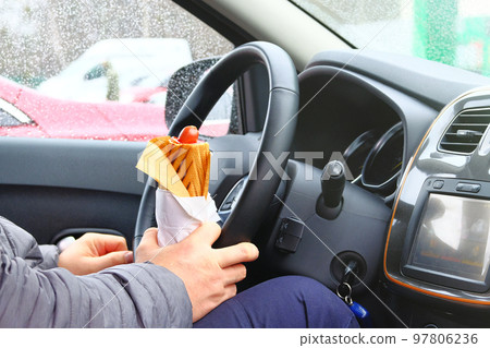 Hot dog in driver hand in his car. Takeaway, social media. Snack in man hand in auto. Hot dog in driver hand in his car. Takeaway, social media. Snack in man hand in auto. 97806236