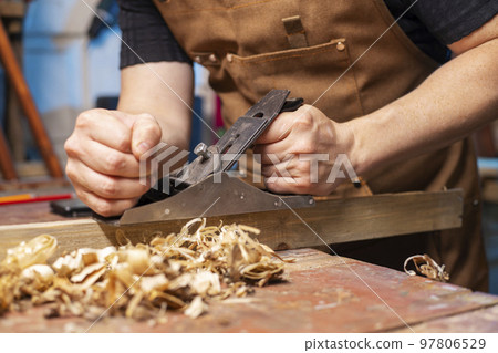 Carpenter in an apron planing a wooden board with a planer, workplace 97806529
