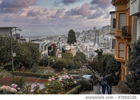 San Francisco, USA. September 20, 2022. Rear view of couple with arms around walking on hilly footpath with cityscape and cloudy sky in the background at sunset San Francisco, USA. September 20, 2022. Rear view of couple with arms around walking on hilly footpath with cityscape and cloudy sky in the background at sunset 97808186