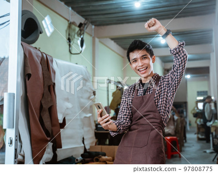 excited asian craftsman smiling using smartphone at leather craft workshop excited asian craftsman smiling using smartphone at leather craft workshop 97808775