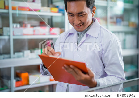 Asian male pharmacist in uniform working with clipboard while registering Asian male pharmacist in uniform working with clipboard while registering 97808942