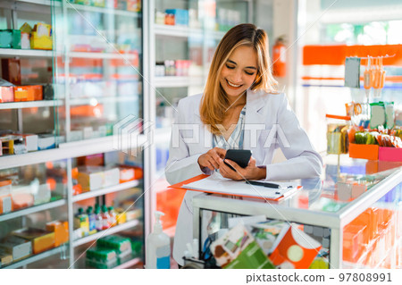 Attractive pharmacist in uniform using smartphone while working 97808991