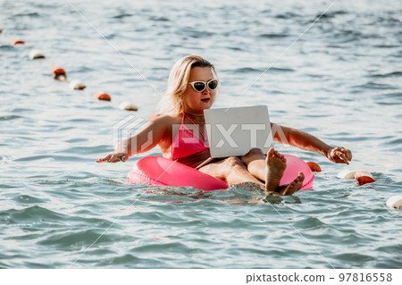 Woman works on laptop in sea. Freelancer, young blond woman in sunglases floating on an inflatable big pink donut with a laptop in the sea at sunset. Freelance, travel and holidays concept. Woman works on laptop in sea. Freelancer, young blond woman in sunglases floating on an inflatable big pink donut with a laptop in the sea at sunset. Freelance, travel and holidays concept. 97816558