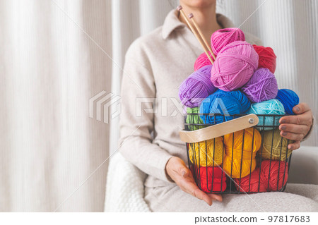 A young woman holds a stylish black wire basket in her arms. Balls and skeins of colored yarn.  97817683