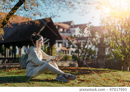 Young Fashionable Teenage Girl With Smartphone In Europian Park In Autumn Sitting At Smiling. Trendy Young Woman In Fall In Park Texting. Retouched, Vibrant Colors. Beautiful Blonde Teenage Girl 97817993