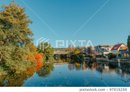 Beautiful small river with clean and clear water front of colorful autumn trees and small old town on the hill agaist nice blue and clouds sky during autumn in Europe Beautiful small river with clean and clear water front of colorful autumn trees and small old town on the hill agaist nice blue and clouds sky during autumn in Europe 97818288
