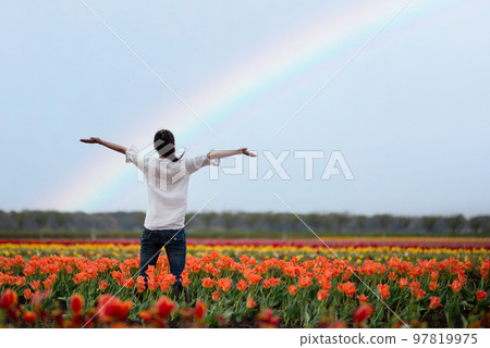 A woman spreading her hands in a rainbow-filled flower field 97819975