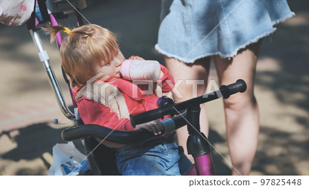 The baby drinks from a bottle while sitting in the pram. 97825448