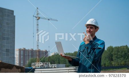 A male builder in a helmet stands against the background of a construction site with a laptop in his hands. A male builder in a helmet stands against the background of a construction site with a laptop in his hands. 97828370