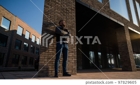Caucasian bearded man in a bomber jacket leaned against a brick wall.  97829605