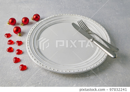 An empty white ceramic plate with cutlery surrounded by red christmas balls and hearts on a light concrete table An empty white ceramic plate with cutlery surrounded by red christmas balls and hearts on a light concrete table 97832241