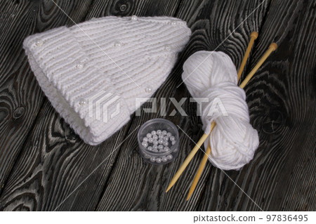 Knitted scarf in white. Women's handmade knitted clothes. Nearby is a skein of white thread for knitting and knitting needles. On black boards. Knitted scarf in white. Women's handmade knitted clothes. Nearby is a skein of white thread for knitting and knitting needles. On black boards. 97836495