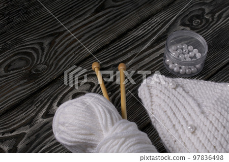 Knitted scarf in white. Women's handmade knitted clothes. Nearby is a skein of white thread for knitting and knitting needles. On black boards. 97836498