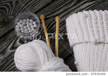Knitted scarf in white. Women's handmade knitted clothes. Nearby is a skein of white thread for knitting and knitting needles. On black boards. Knitted scarf in white. Women's handmade knitted clothes. Nearby is a skein of white thread for knitting and knitting needles. On black boards. 97836499