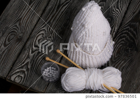 Knitted scarf in white. Women's handmade knitted clothes. Nearby is a skein of white thread for knitting and knitting needles. On black boards. Knitted scarf in white. Women's handmade knitted clothes. Nearby is a skein of white thread for knitting and knitting needles. On black boards. 97836501