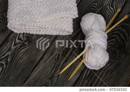 Knitted scarf in white. Women's handmade knitted clothes. Nearby is a skein of white thread for knitting and knitting needles. On black boards. Knitted scarf in white. Women's handmade knitted clothes. Nearby is a skein of white thread for knitting and knitting needles. On black boards. 97836502