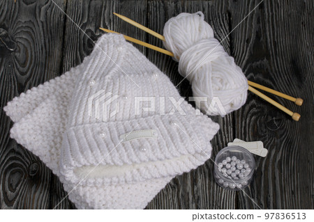 Knitted hat and scarf in white. Women's handmade knitted clothes. Nearby is a skein of white thread for knitting and knitting needles. Beads for decoration in a container. On black boards. Knitted hat and scarf in white. Women's handmade knitted clothes. Nearby is a skein of white thread for knitting and knitting needles. Beads for decoration in a container. On black boards. 97836513