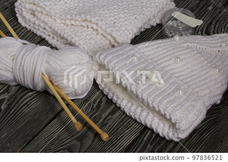 Knitted hat and scarf in white. Women's handmade knitted clothes. Nearby is a skein of white thread for knitting and knitting needles. Beads for decoration in a container. On black boards. Knitted hat and scarf in white. Women's handmade knitted clothes. Nearby is a skein of white thread for knitting and knitting needles. Beads for decoration in a container. On black boards. 97836521