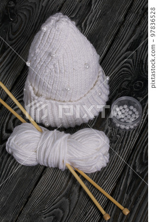 Knitted scarf in white. Women's handmade knitted clothes. Nearby is a skein of white thread for knitting and knitting needles. On black boards. Knitted scarf in white. Women's handmade knitted clothes. Nearby is a skein of white thread for knitting and knitting needles. On black boards. 97836528
