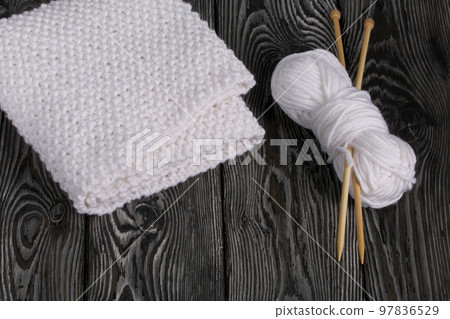 Knitted scarf in white. Women's handmade knitted clothes. Nearby is a skein of white thread for knitting and knitting needles. On black boards. Knitted scarf in white. Women's handmade knitted clothes. Nearby is a skein of white thread for knitting and knitting needles. On black boards. 97836529