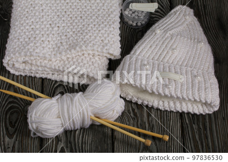 Knitted hat and scarf in white. Women's handmade knitted clothes. Nearby is a skein of white thread for knitting and knitting needles. Beads for decoration in a container. On black boards. Knitted hat and scarf in white. Women's handmade knitted clothes. Nearby is a skein of white thread for knitting and knitting needles. Beads for decoration in a container. On black boards. 97836530