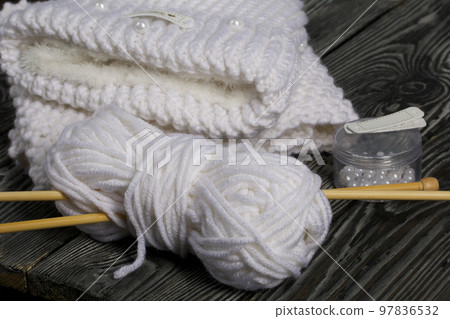 Knitted hat and scarf in white. Women's handmade knitted clothes. Nearby is a skein of white thread for knitting and knitting needles. Beads for decoration in a container. On black boards. Knitted hat and scarf in white. Women's handmade knitted clothes. Nearby is a skein of white thread for knitting and knitting needles. Beads for decoration in a container. On black boards. 97836532