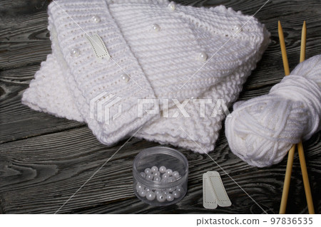 Knitted hat and scarf in white. Women's handmade knitted clothes. Nearby is a skein of white thread for knitting and knitting needles. Beads for decoration in a container. On black boards. 97836535