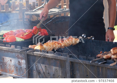 Chef frying meat and vegetables on grill. Man in black apron roasted red peppers and zucchini. Barbecue in open air. Chef frying meat and vegetables on grill. Man in black apron roasted red peppers and zucchini. Barbecue in open air. 97837408
