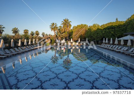 Empty loungers and folded parasols surrounded with trees arranged at poolside with clear blue sky in the background at tourist resort 97837718
