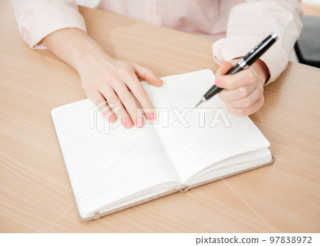 Close up of a woman's hands writing in a notepad placed on a wooden table, home office and work concept, plans and forecasts, left-handed Close up of a woman's hands writing in a notepad placed on a wooden table, home office and work concept, plans and forecasts, left-handed 97838972