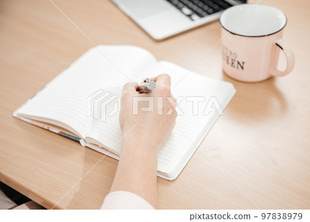 Close up of a woman's hands writing in a notepad placed on a wooden table, laptop, home office and work concept, plans and thoughts Close up of a woman's hands writing in a notepad placed on a wooden table, laptop, home office and work concept, plans and thoughts 97838979