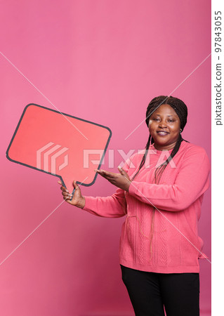 Joyful smiling adult woman holding blank template announcement placard posing in studio. African american confident woman holding red cardboard speech bubble on pink background. Joyful smiling adult woman holding blank template announcement placard posing in studio. African american confident woman holding red cardboard speech bubble on pink background. 97843055