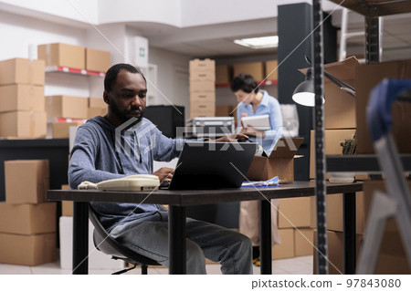 African american employee sitting at desk in warehouse delivery department looking at cargo stock on laptop before start preparing helmet order. Storehouse worker using carton box and bubble wrap 97843080